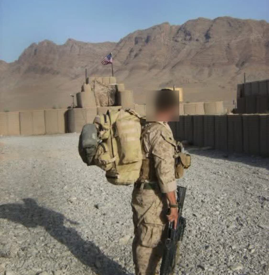 US Marine carrying a pack at a forward post, American flag flying over the barriers